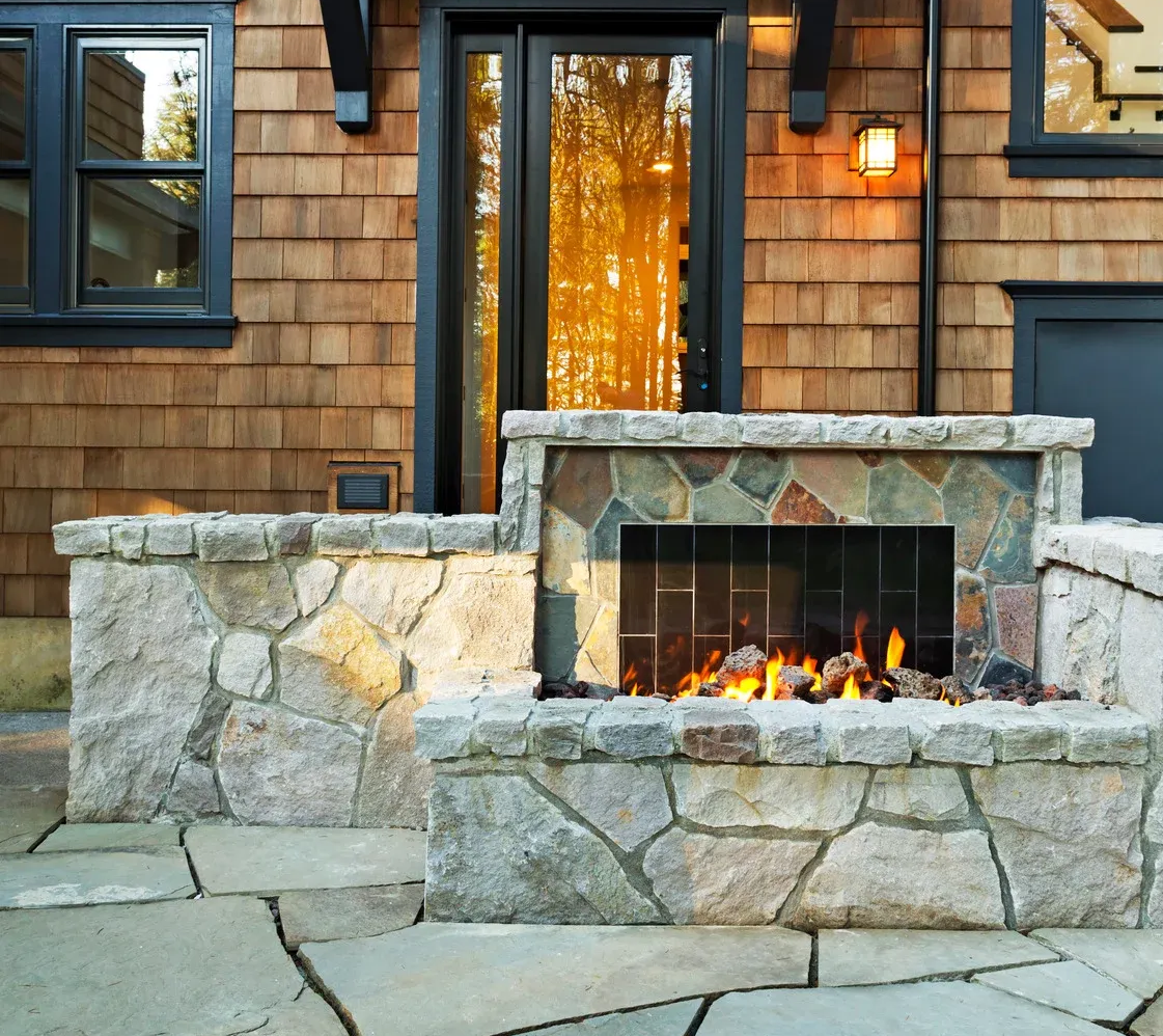 Stone patio with a small outdoor fireplace beneath a dark shingled house facade and lit doorway