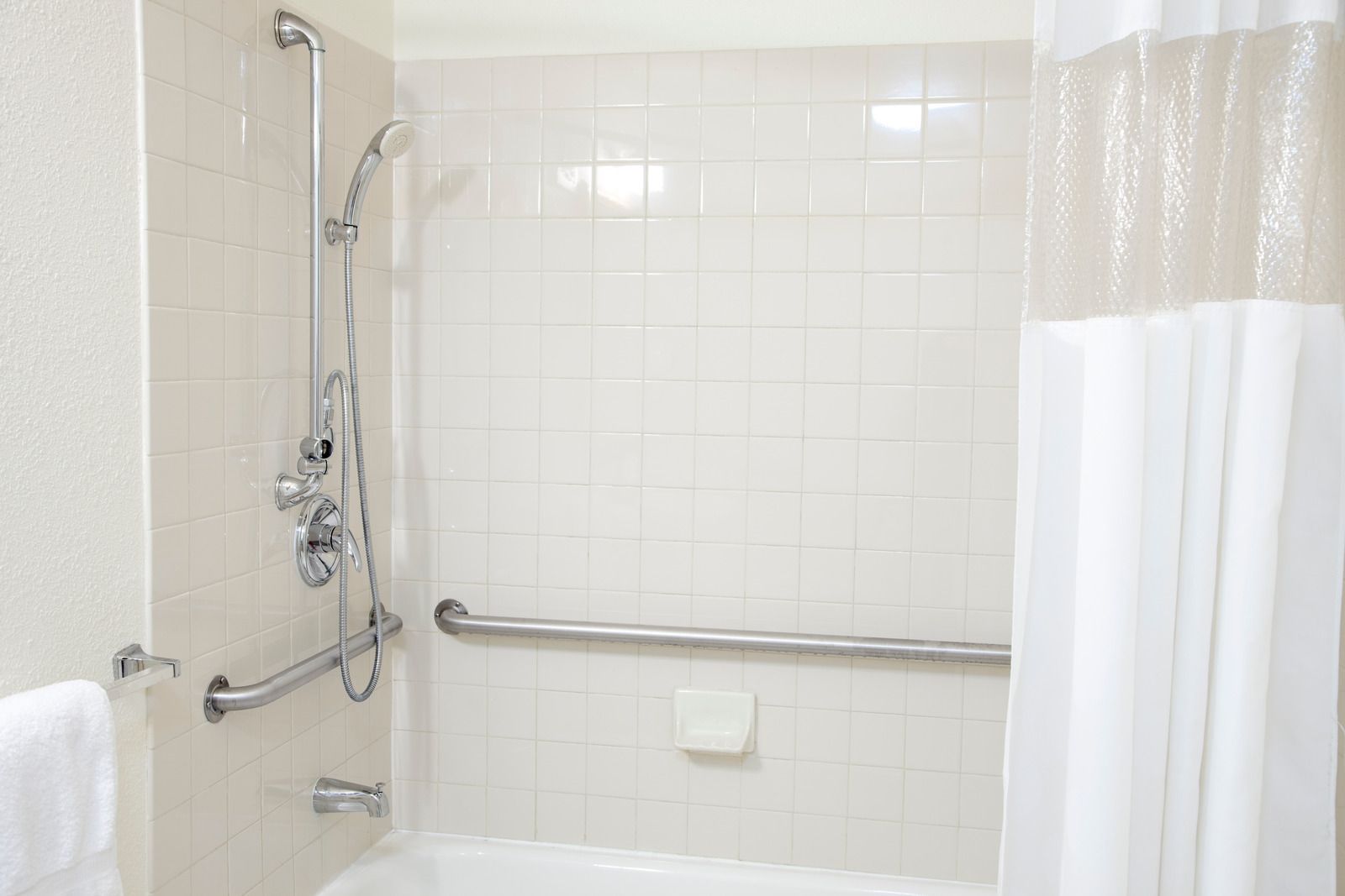 White tiled bathroom with walk-in shower, grab bars, handheld showerhead, and partially closed curtain