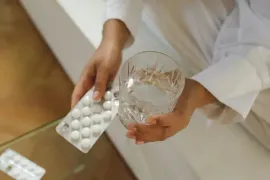 Hands holding a blister pack of white pills and a glass of water on a couch