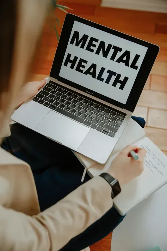 Laptop displaying “MENTAL HEALTH” on a person’s lap while they write in a notebook