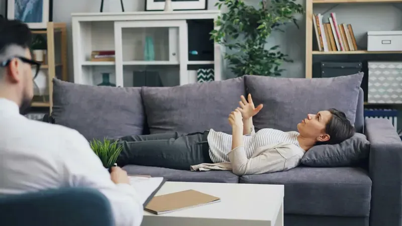 Two people talking in a bright living room, one reclining on a gray sofa with a tablet