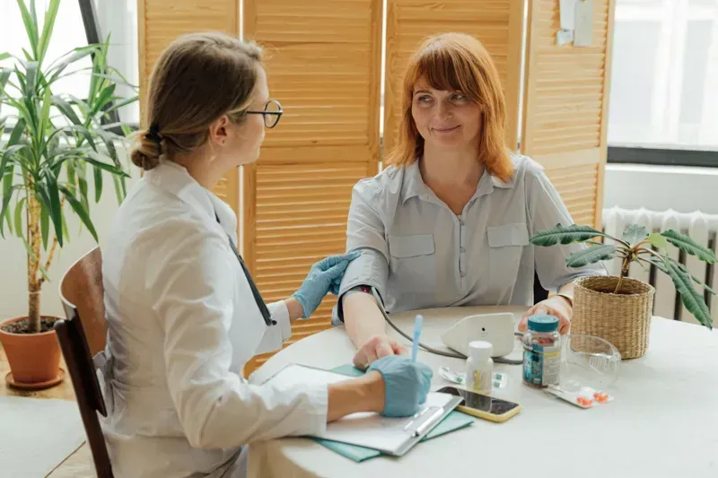 Two women in a bright clinic room, one taking the other’s blood pressure at a table.