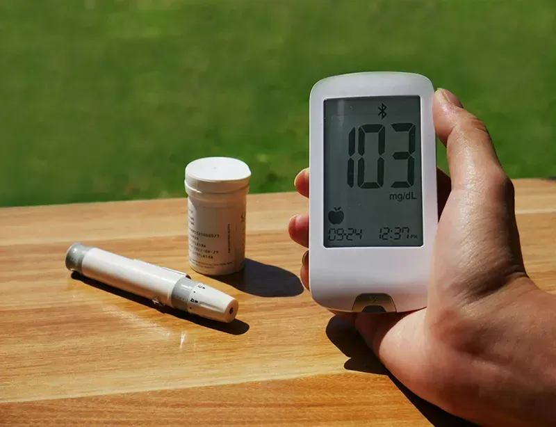 Hand holding a white digital meter reading 103 on a wooden table with a test strip and container outdoors