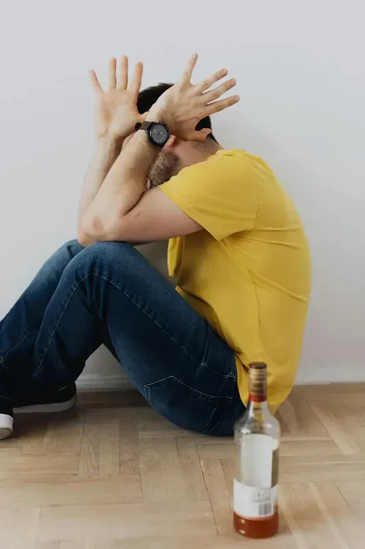 Person in a yellow shirt sits on the floor with hands over face beside a bottle of alcohol.