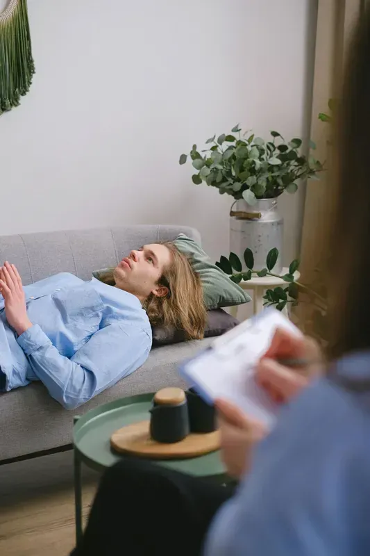Woman lying on a sofa in a counseling session, holding a book, with a therapist taking notes nearby