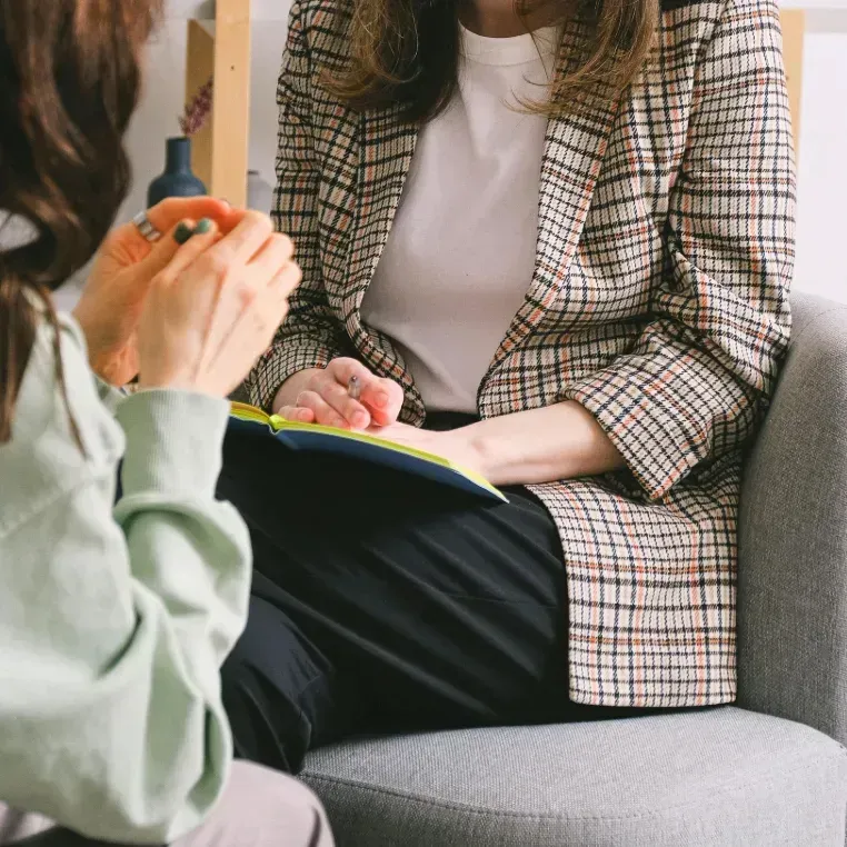 Two people talking in a gray armchair, one holding a notebook in a plaid blazer.