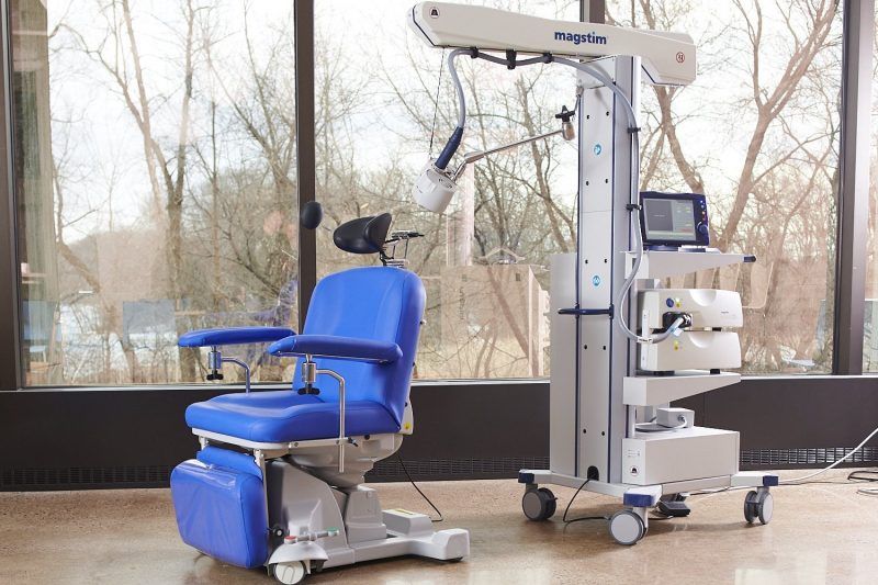 Blue medical chair beside a wheeled TMS therapy machine in a bright clinic room
