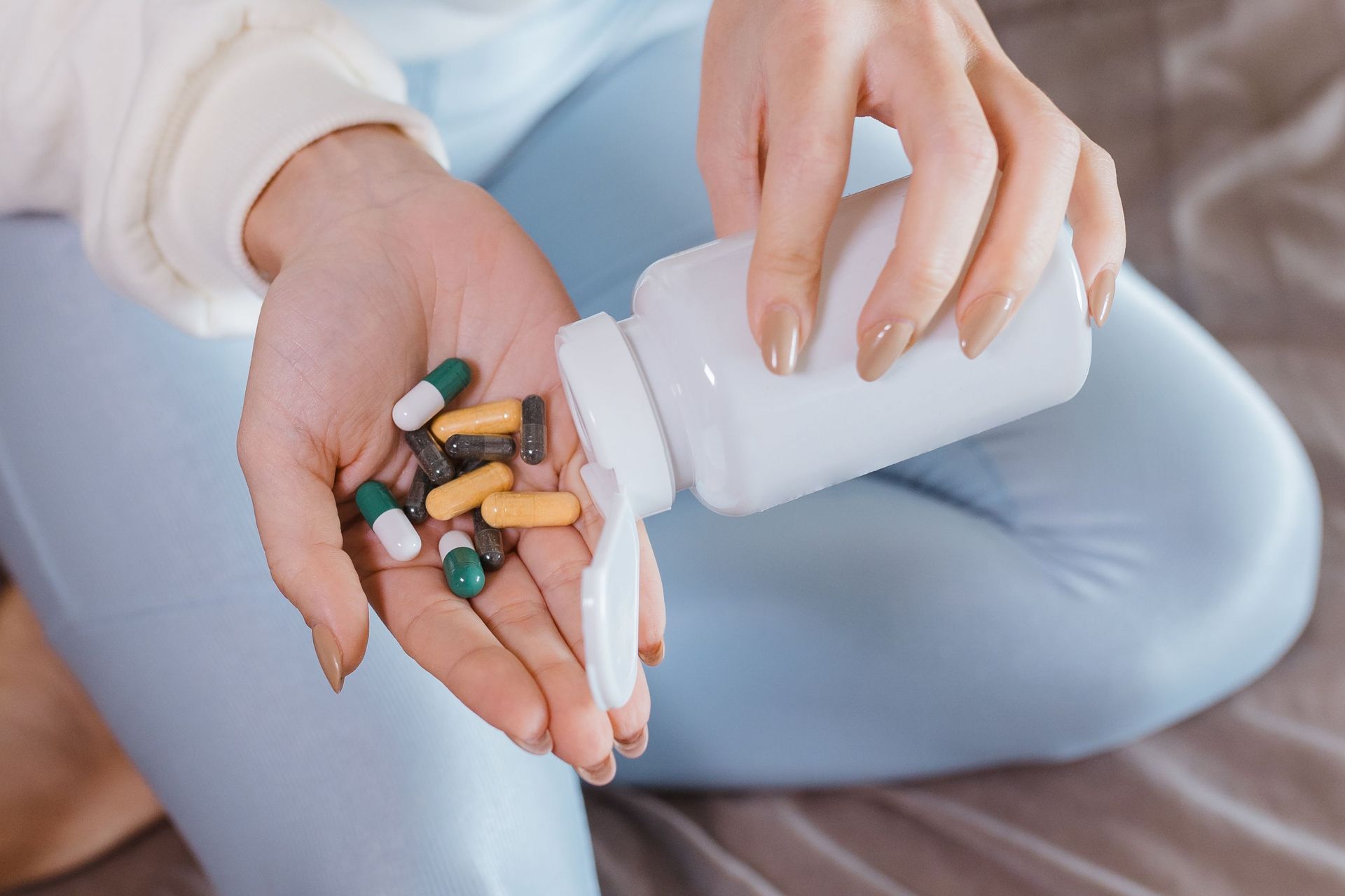 Hands pouring assorted pills from a white bottle onto a palm on a bed