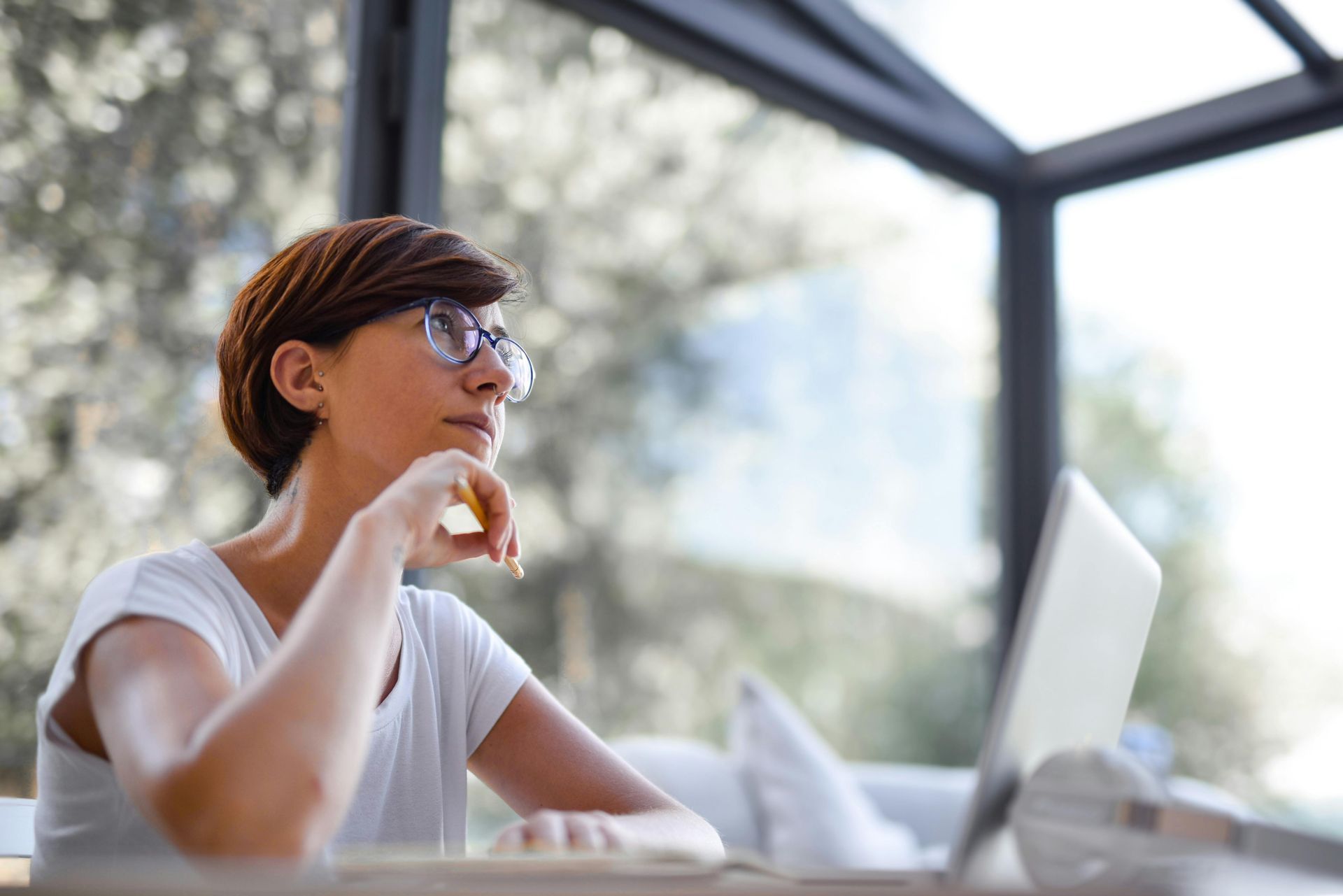 Person sitting outdoors with a laptop, holding a drink and looking out through a patio window.
