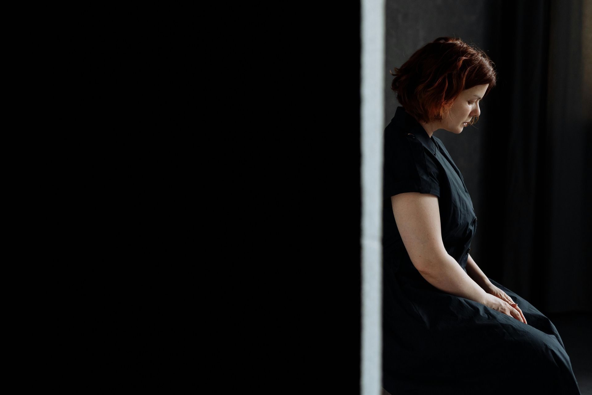 Woman in dark dress sits, head down, against a wall. The setting appears somber, with a black background.