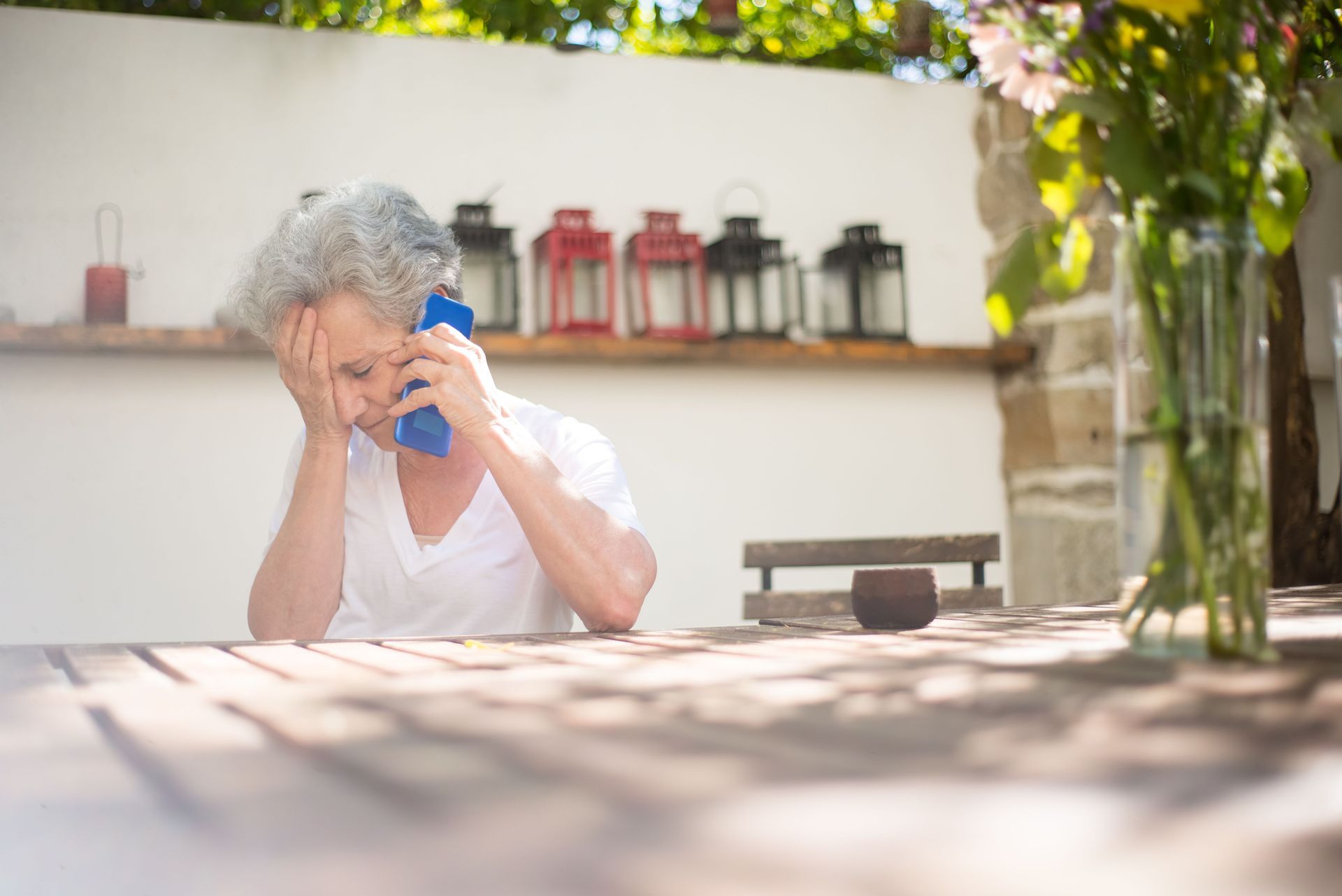 Person in a white shirt sitting outdoors, wiping their face with a blue cloth at a sunlit table.