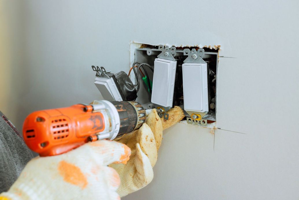 Hand drilling a hole in a wall beside three electrical outlets with exposed wiring