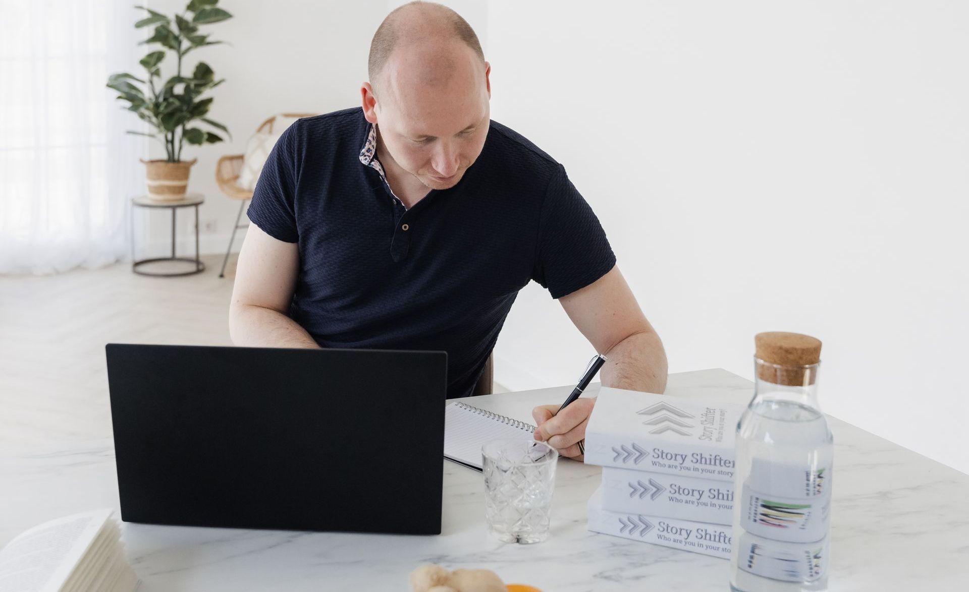 Een man werkt aan een laptop en schrijft op papier. Op de achtergrond staan een witte tafel, een waterfles en een potplant.