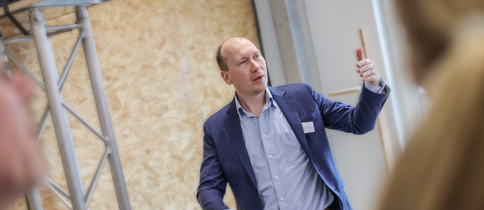 Een man in een blauwe blazer en een wit overhemd gebaart met een pen. Hij staat in een kamer met een lichtgekleurde muur.