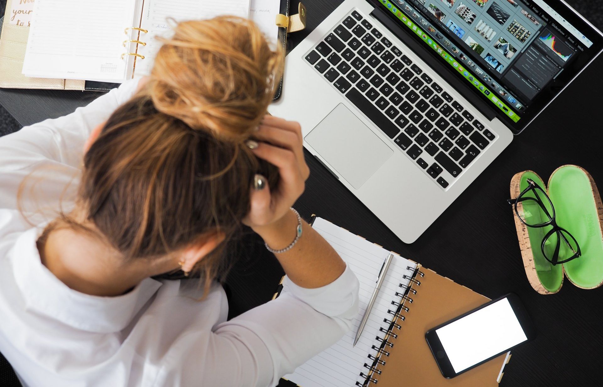 Een vrouw zit gestrest achter haar bureau, met haar hoofd in haar handen, en heeft een laptop, notitieboekje, telefoon en bril bij zich.