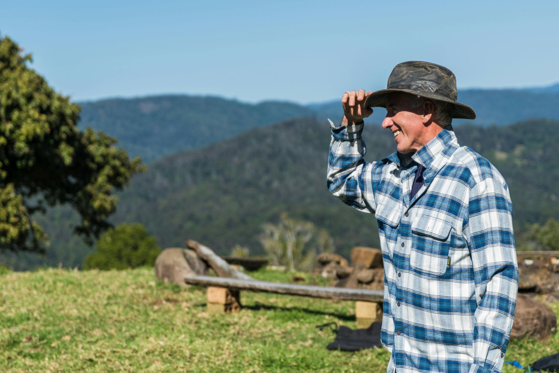 Man in plaid shirt and hat, shielding eyes, looking at mountains in the distance.