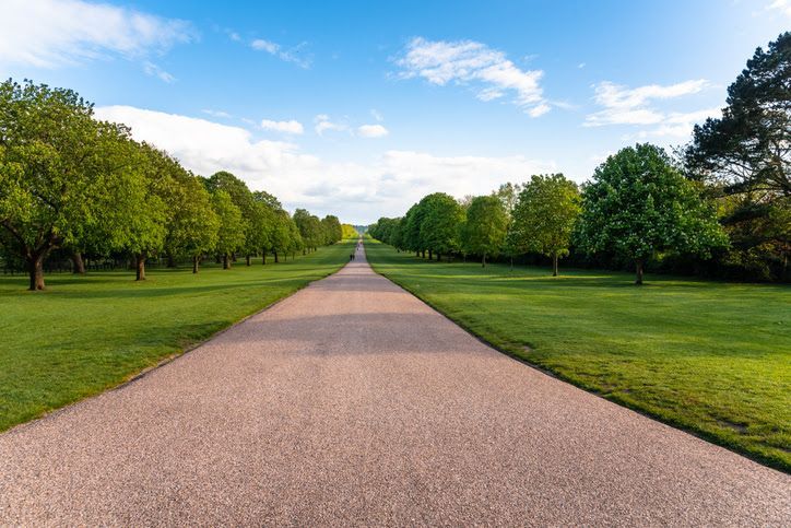 A long, straight gravel path stretches into the distance, flanked by lush green lawns and rows of mature trees under a blue sky.