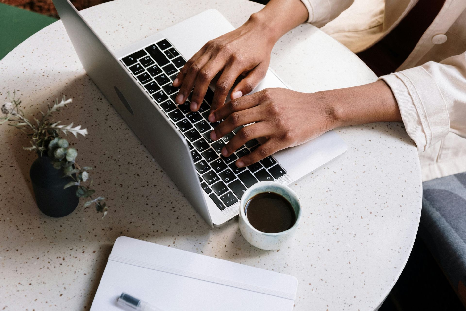 Hands typing on a laptop at a table with a coffee cup and small plant.