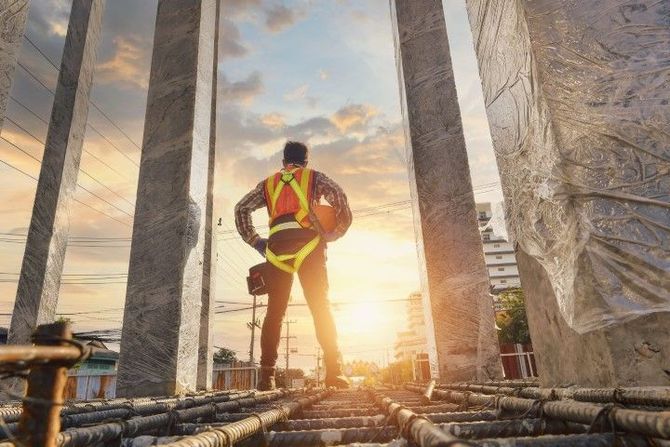 Construction worker in safety vest and harness standing on scaffolding at sunset, looking toward the horizon
