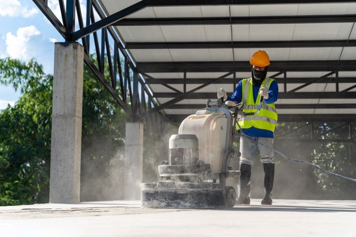 Worker in a hard hat using a floor grinder in a concrete structure, with dust rising.
