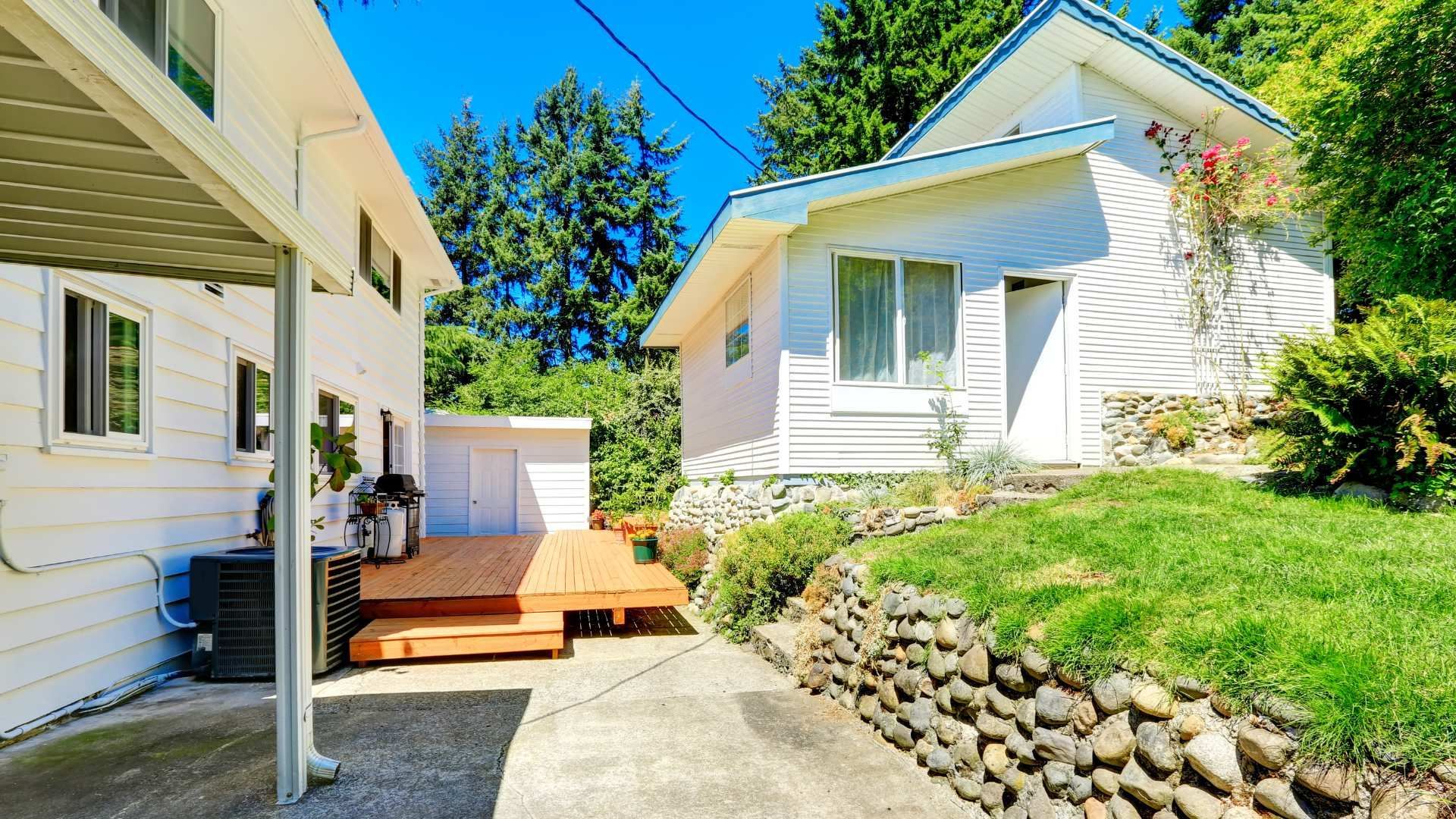 White house exterior with small wooden deck, concrete path, and stone retaining wall in a sunny yard