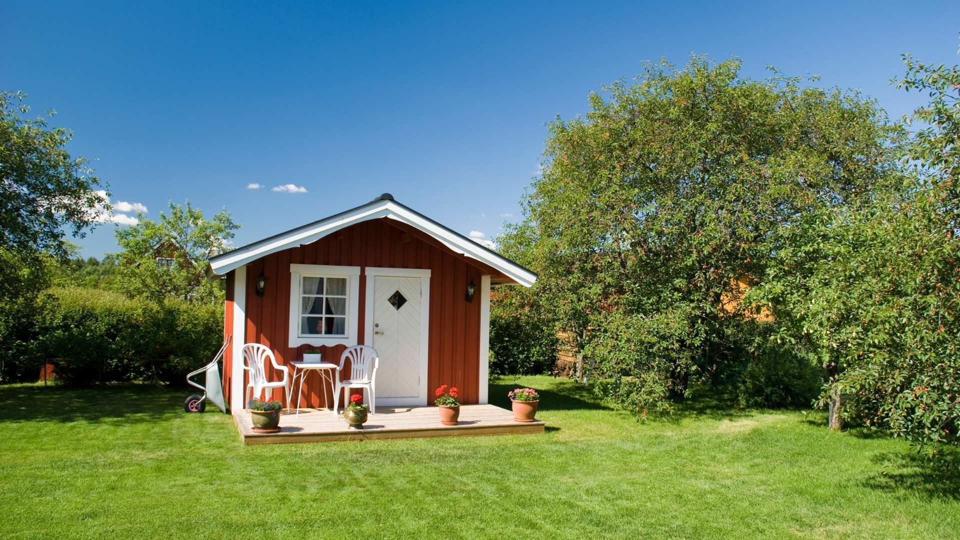 Small red cottage with a porch and chairs in a green garden under a blue sky