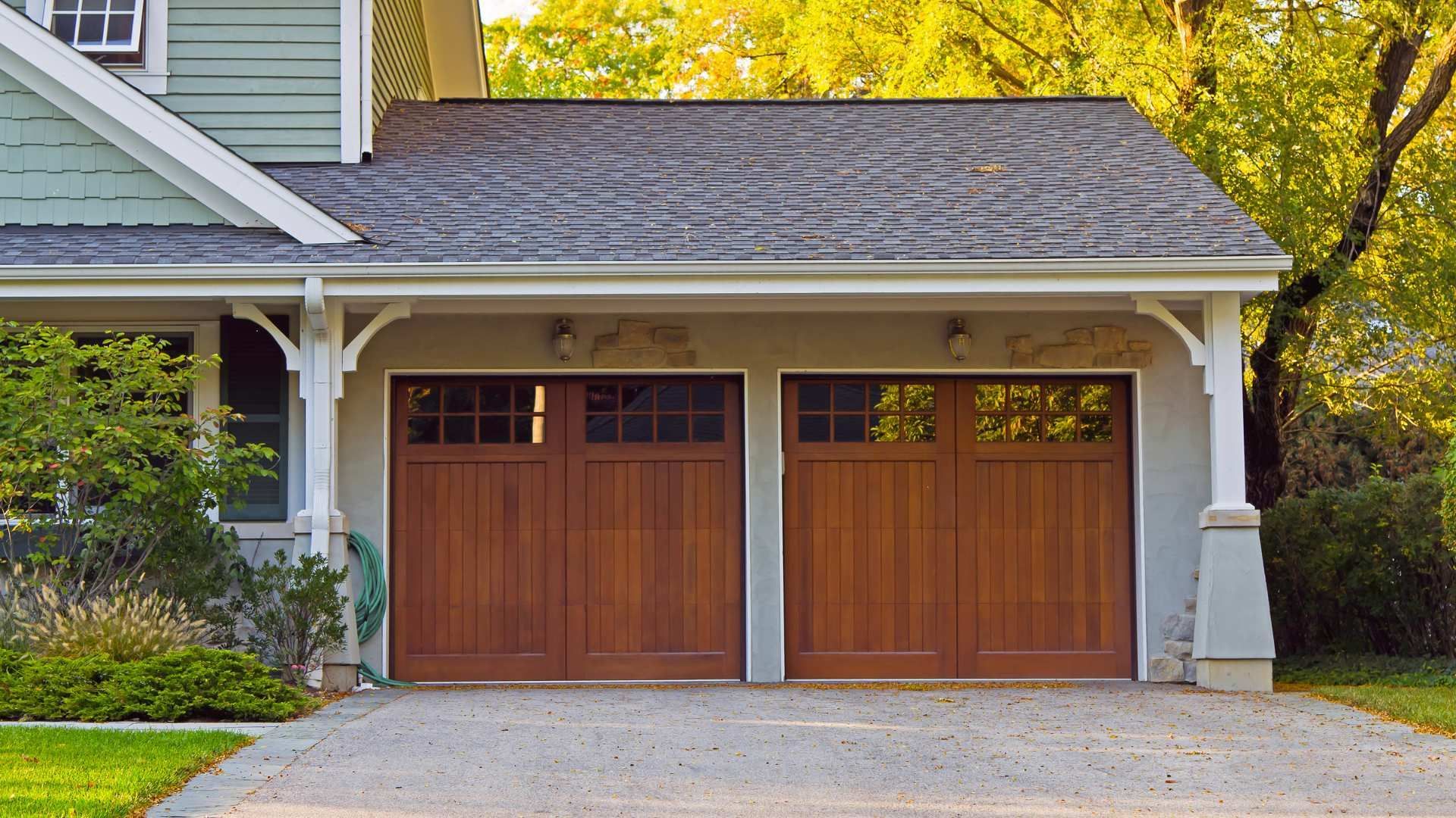 Two wooden garage doors on a house with gray siding and a tree overhead
