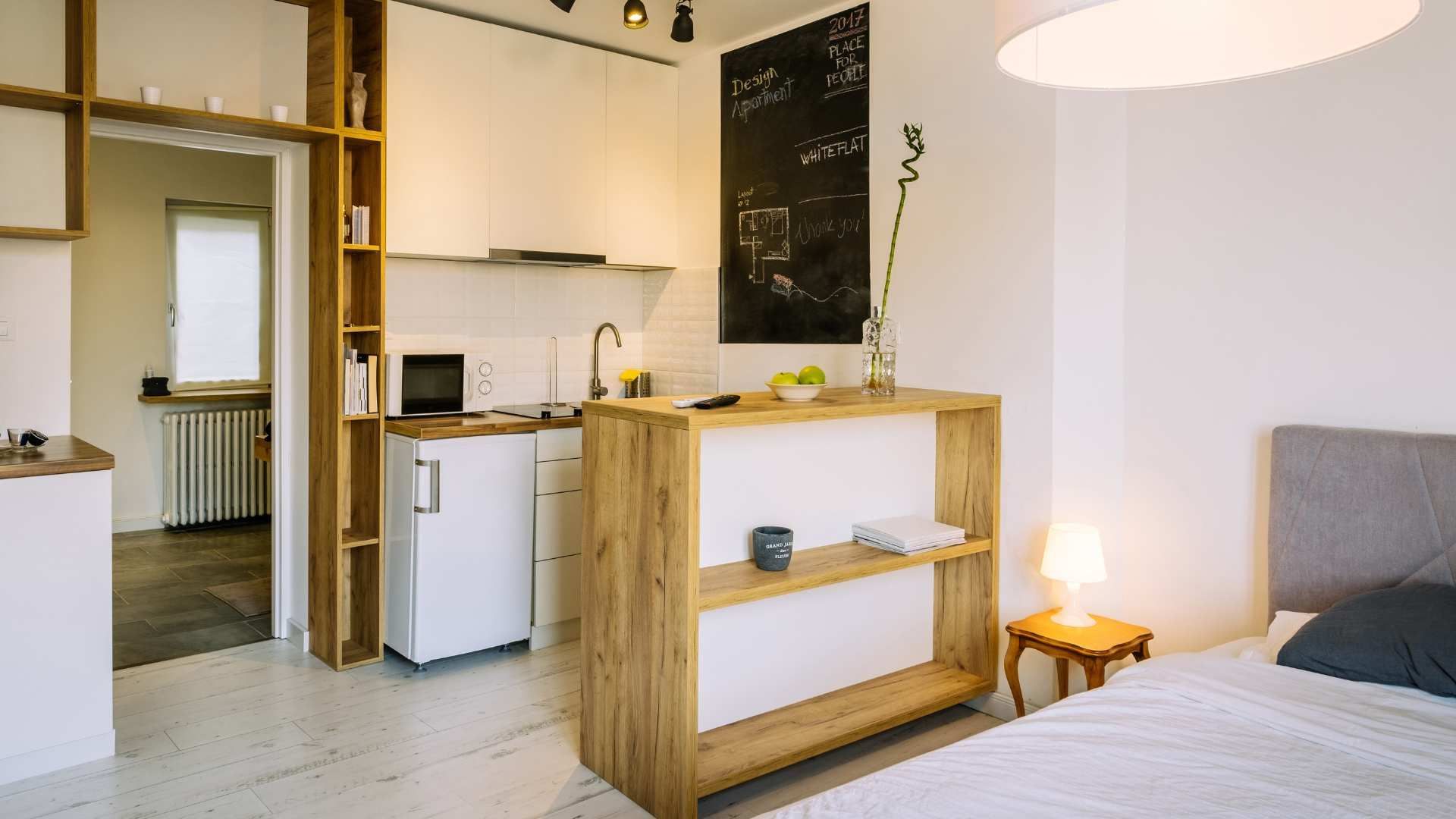 Small studio apartment with kitchenette, wooden shelf, and bed by a bedside lamp