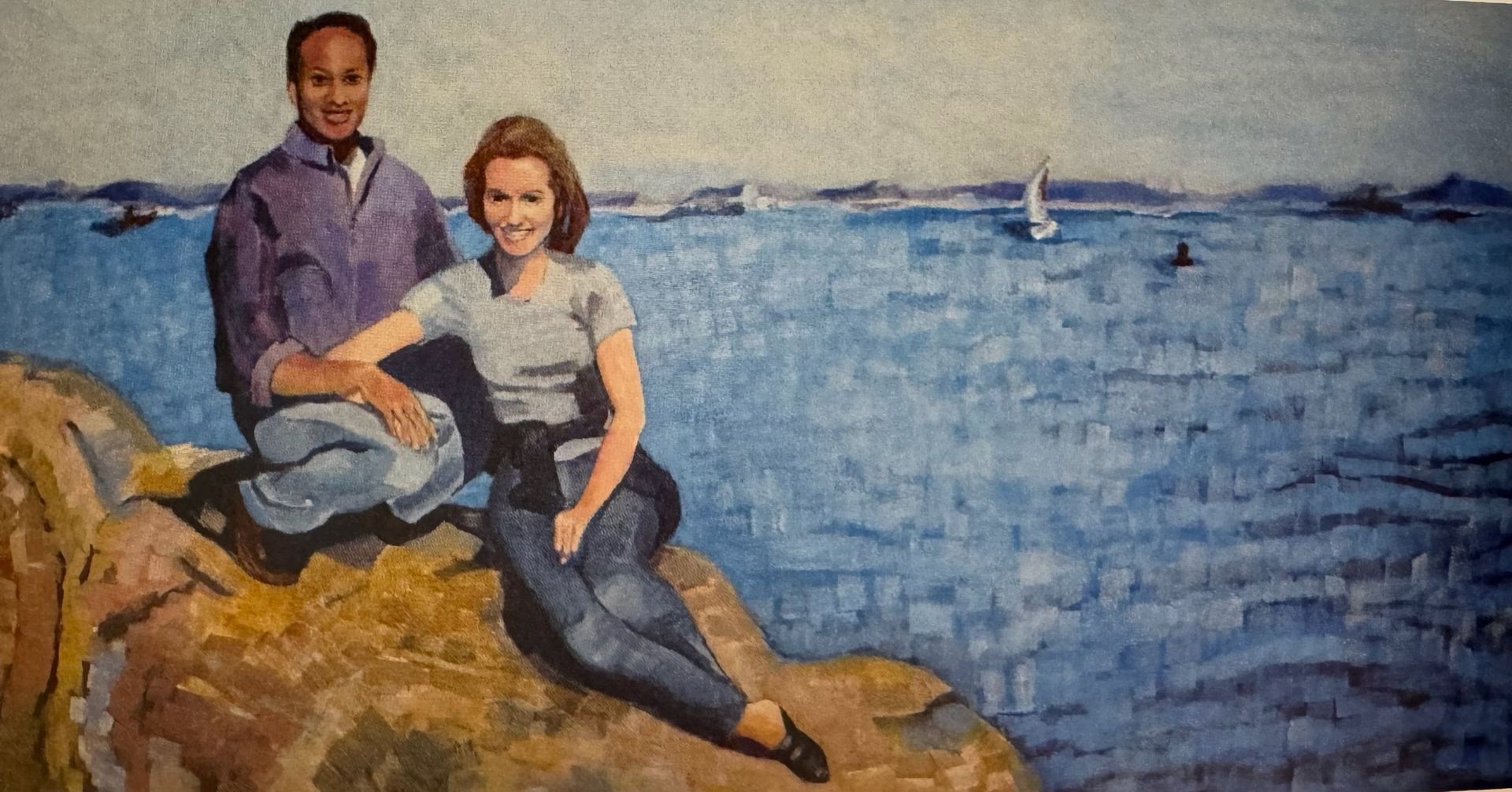 Two people sitting on rocks by a blue waterfront, smiling at the camera