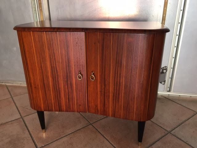 Piccolo mobiletto in legno in stile mid-century, con due ante e gambe nere affusolate, su un pavimento piastrellato.