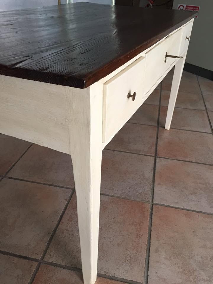 Tavolo in legno bianco con piano scuro, visto di tre quarti su un pavimento piastrellato.