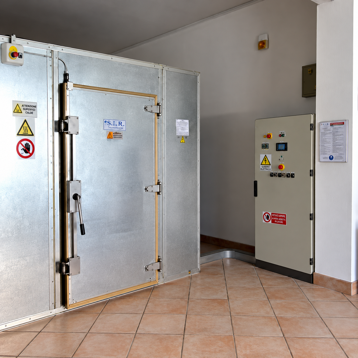 Cella frigorifera industriale aperta, con una grande porta metallica e un pannello di controllo in un corridoio piastrellato.