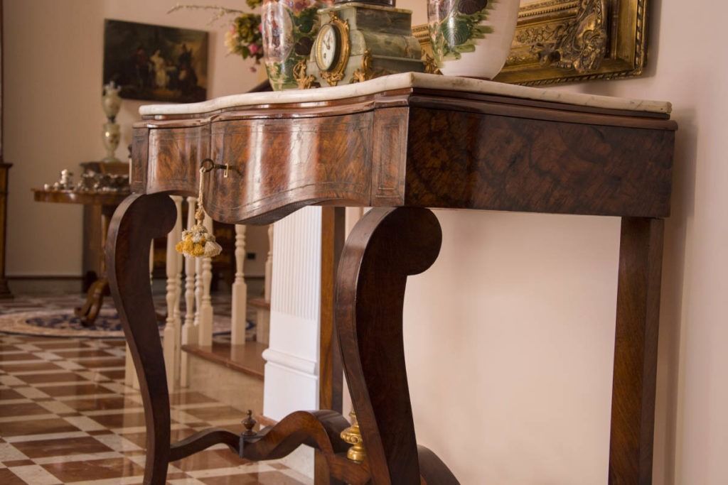Consolle antica in legno con gambe ornate in un corridoio, decorata con fiori e oggetti ornamentali.