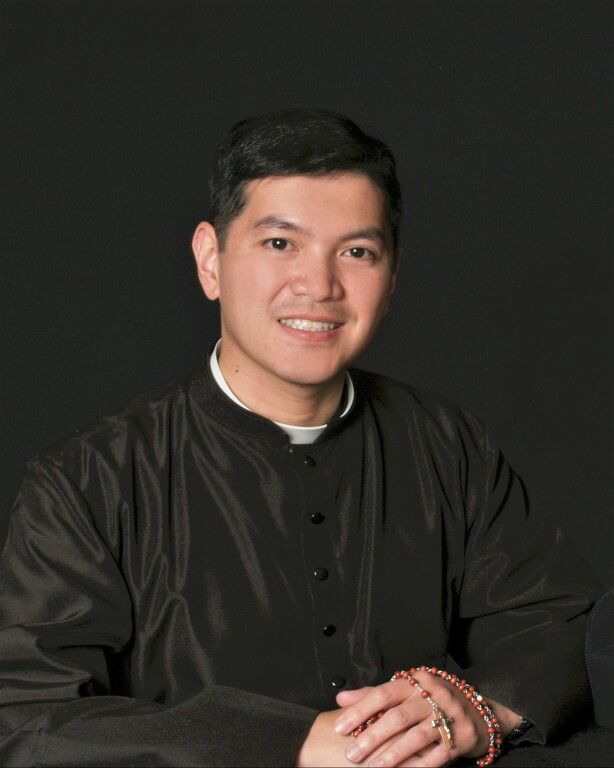 Smiling person in black clerical clothing with rosary, seated against a dark background