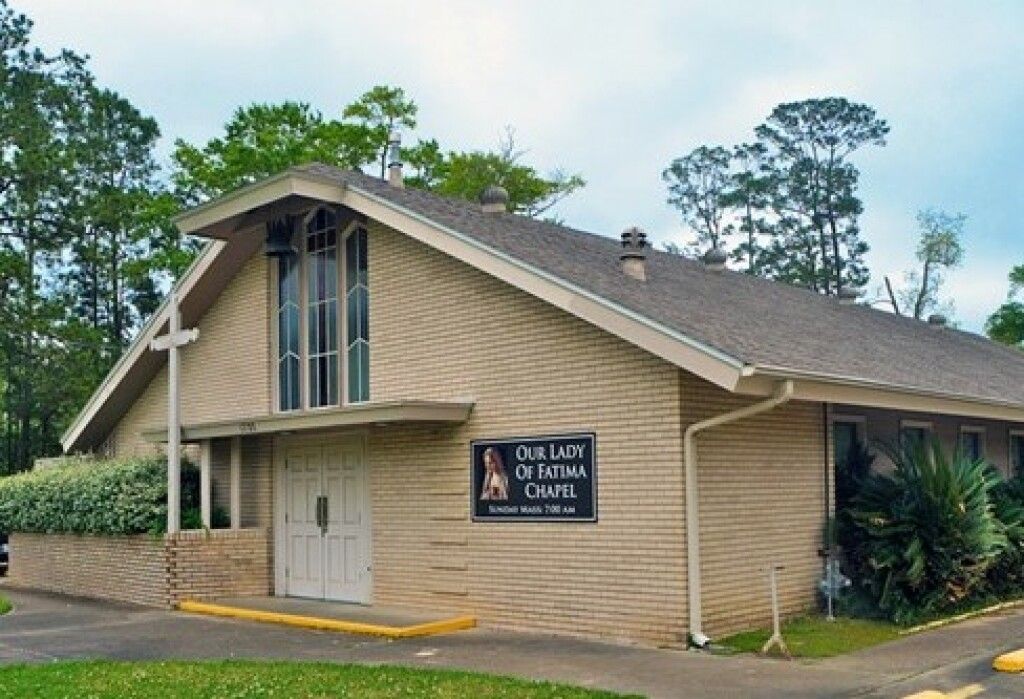 Small beige church or chapel with a sign, surrounded by trees and greenery under a cloudy sky