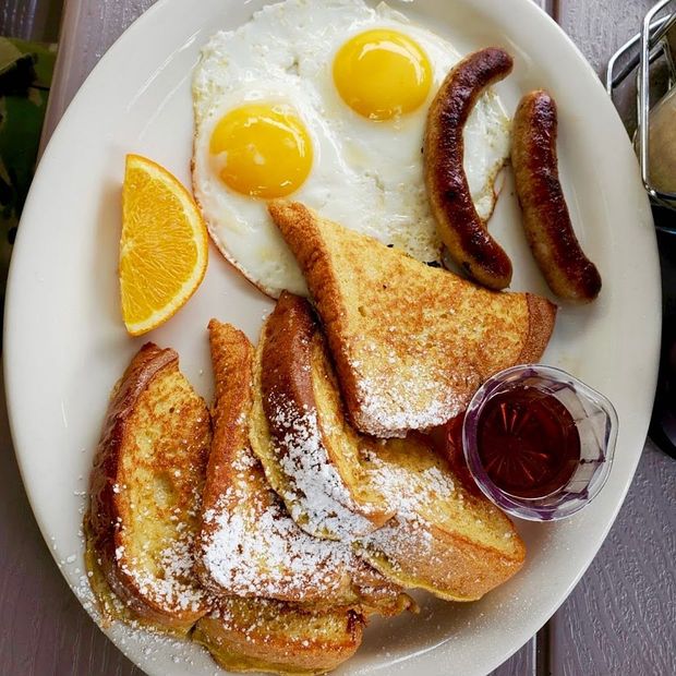 Breakfast plate with French toast, two sunny-side-up eggs, sausages, an orange slice, and syrup.