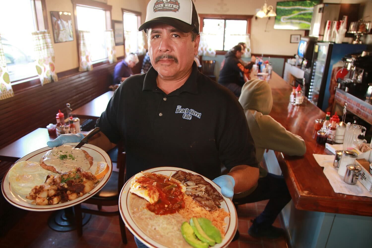 Waiter in a restaurant holding two plated meals, with diners and booths in the background.