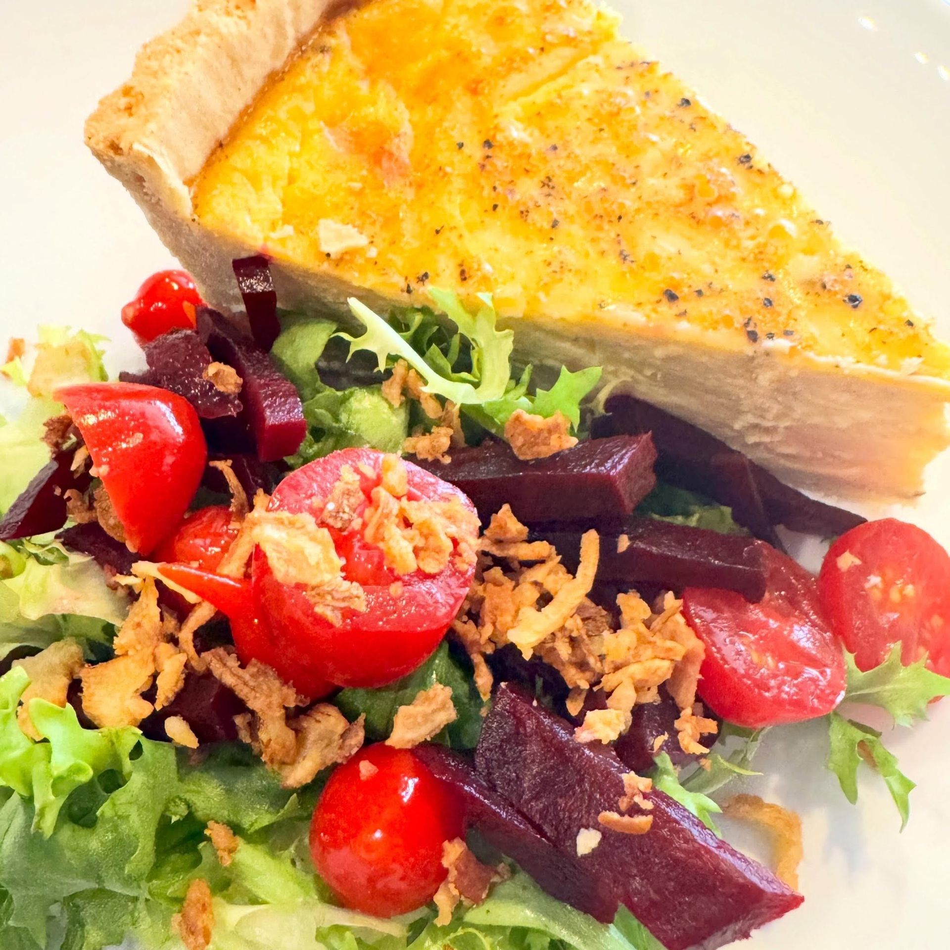 Quiche slice with mixed green salad, cherry tomatoes, and beet pieces on a white plate