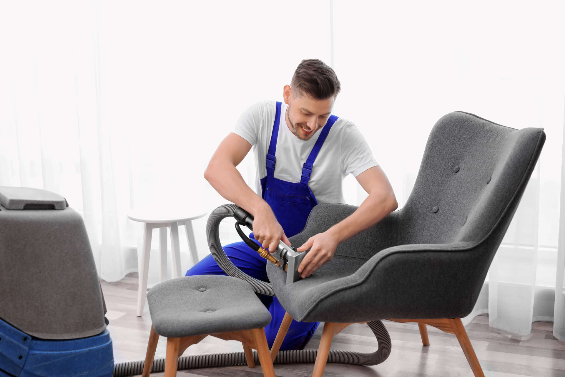 Man cleaning a gray armchair with a vacuum cleaner in a bright room.