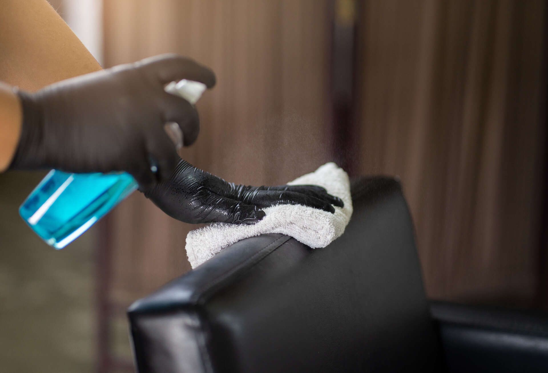 Person wearing black gloves sprays and wipes a black chair with a blue liquid and white cloth.