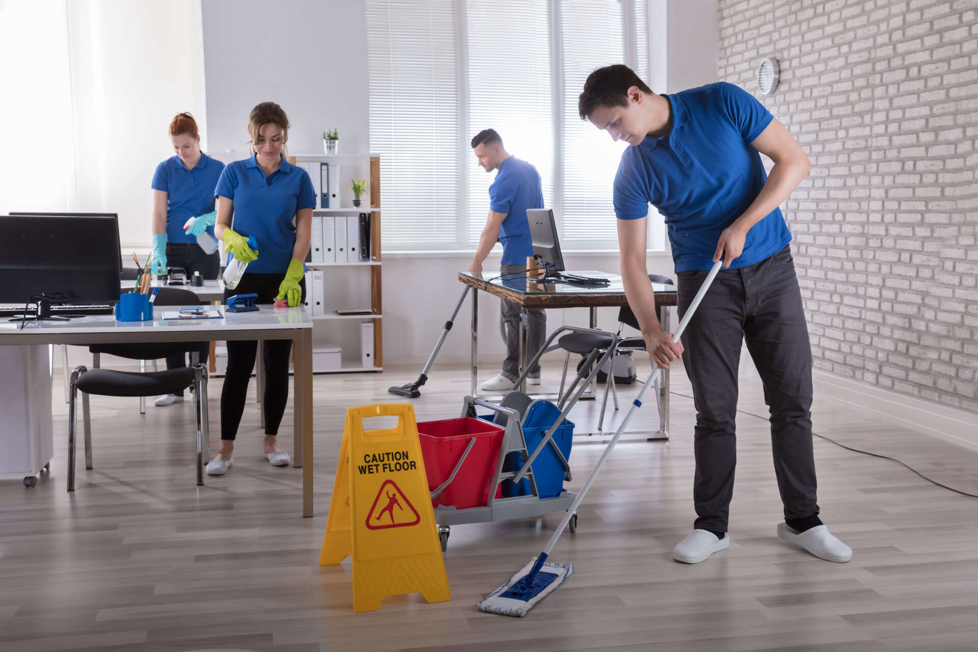 Office cleaning crew in blue shirts cleans a workspace: mopping, vacuuming, dusting.