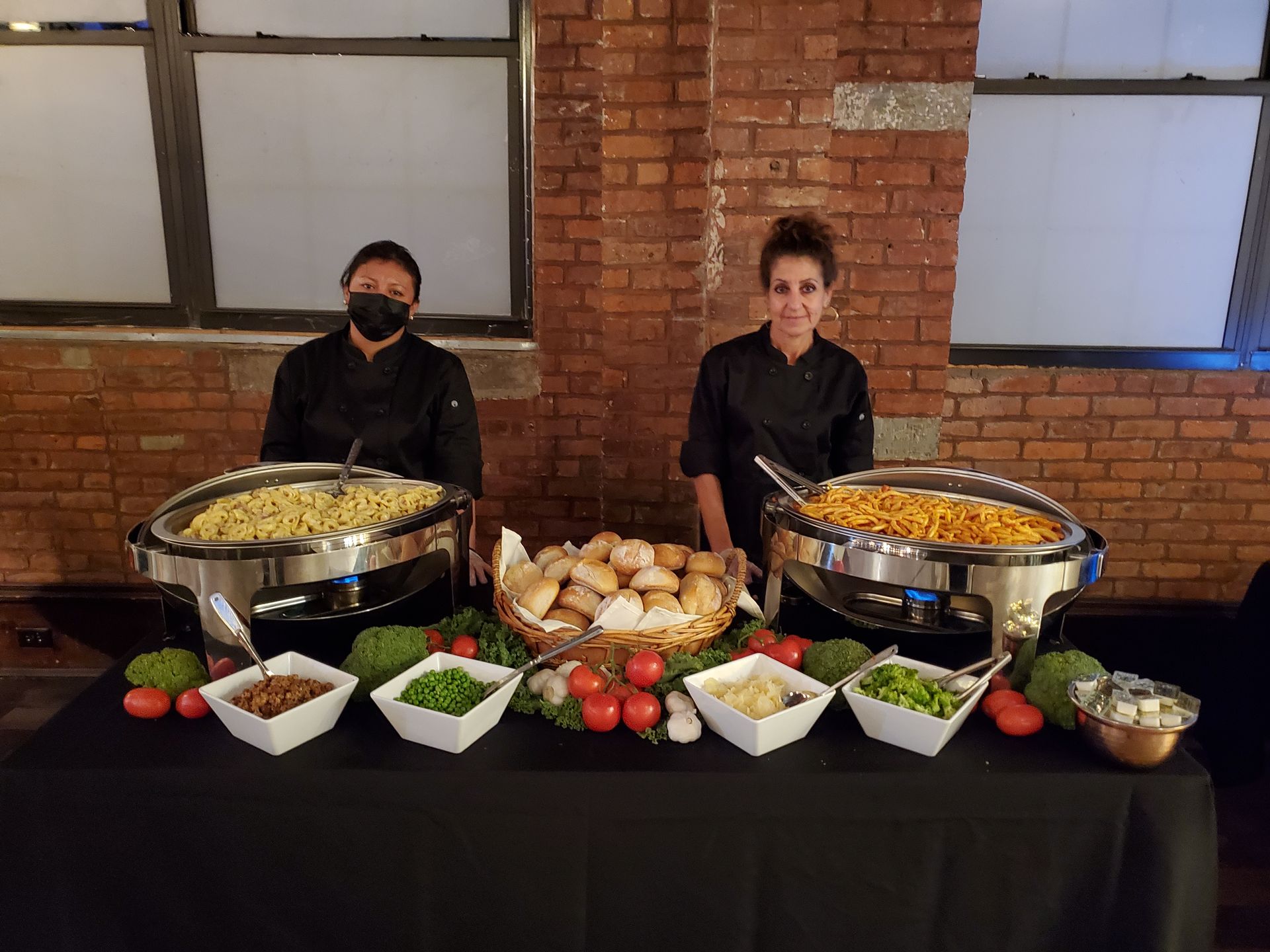 Two catering staff stand behind a buffet table featuring chafing dishes, a basket of rolls, and bowls of side dishes.