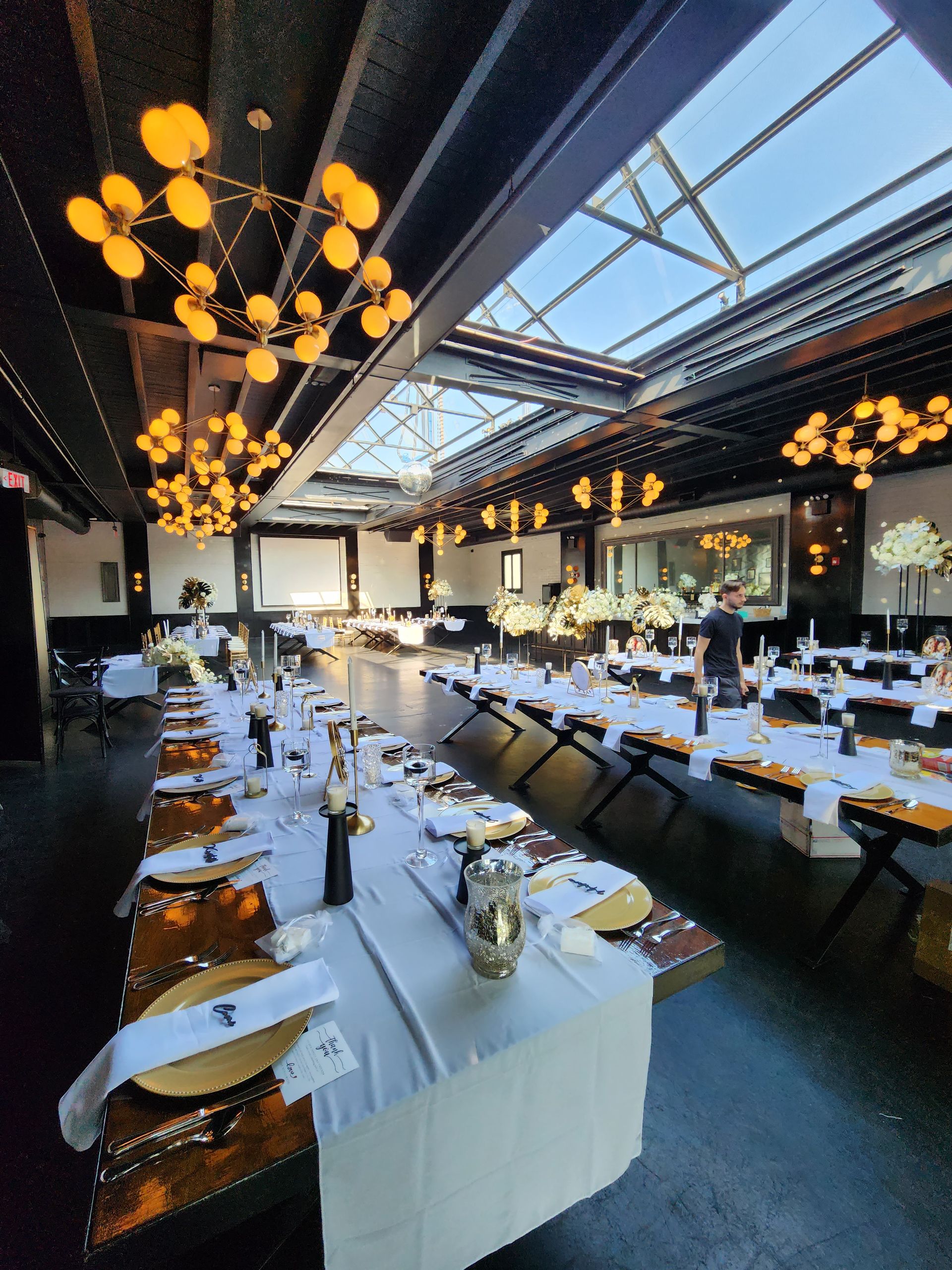 A long, elegant dining table set for an event in a spacious venue with modern globe chandeliers and a glass roof.
