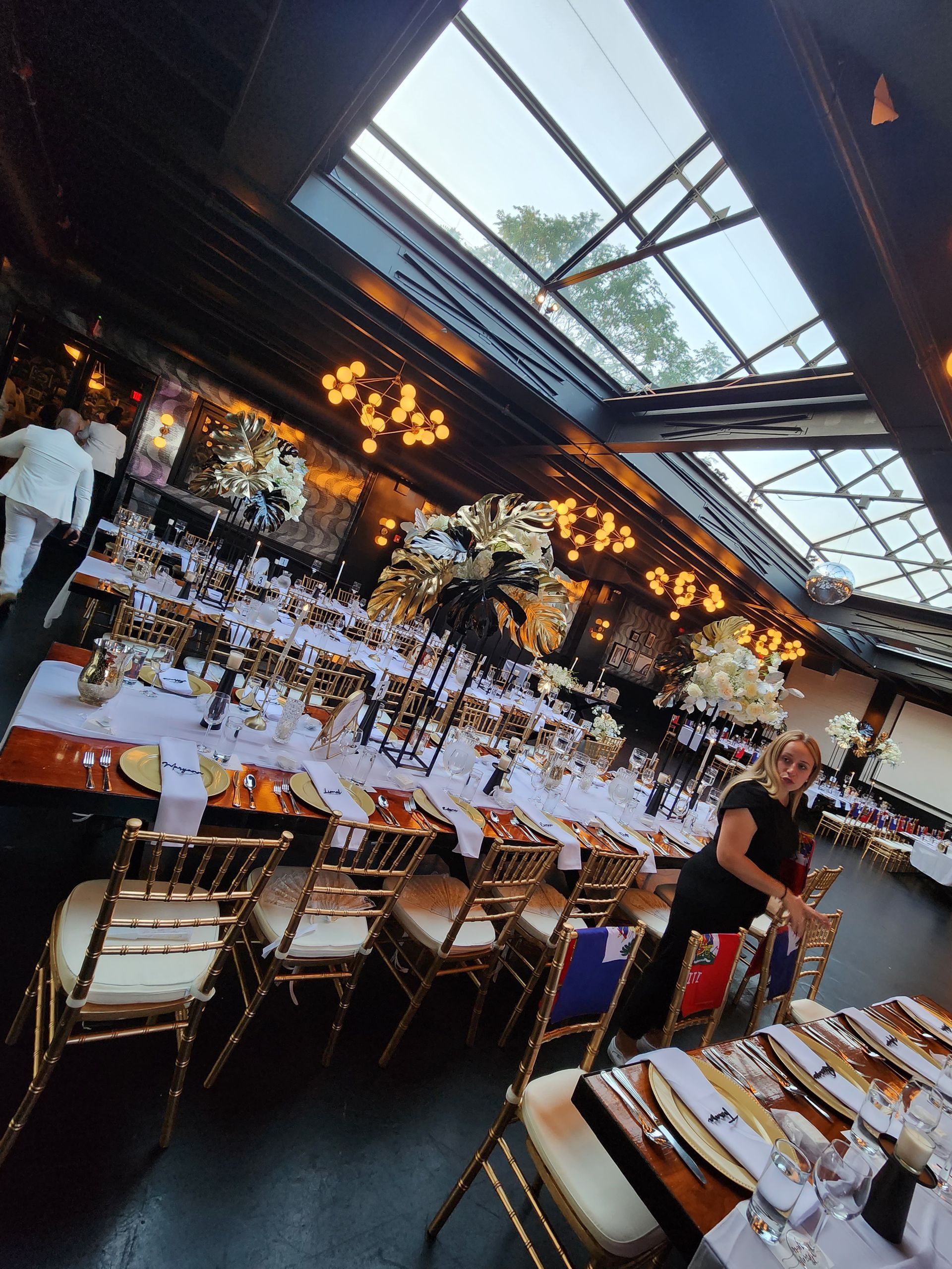 An event space with long dining tables set with gold chairs, elegant centerpieces, and a large skylight above.