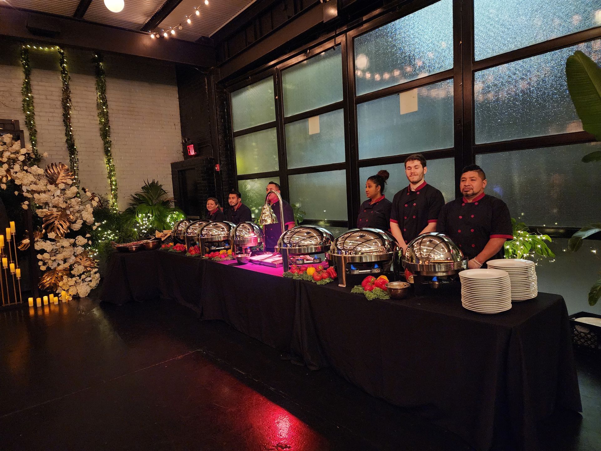 Catering staff stand behind a long buffet table with silver chafing dishes and stacks of plates in an event space.