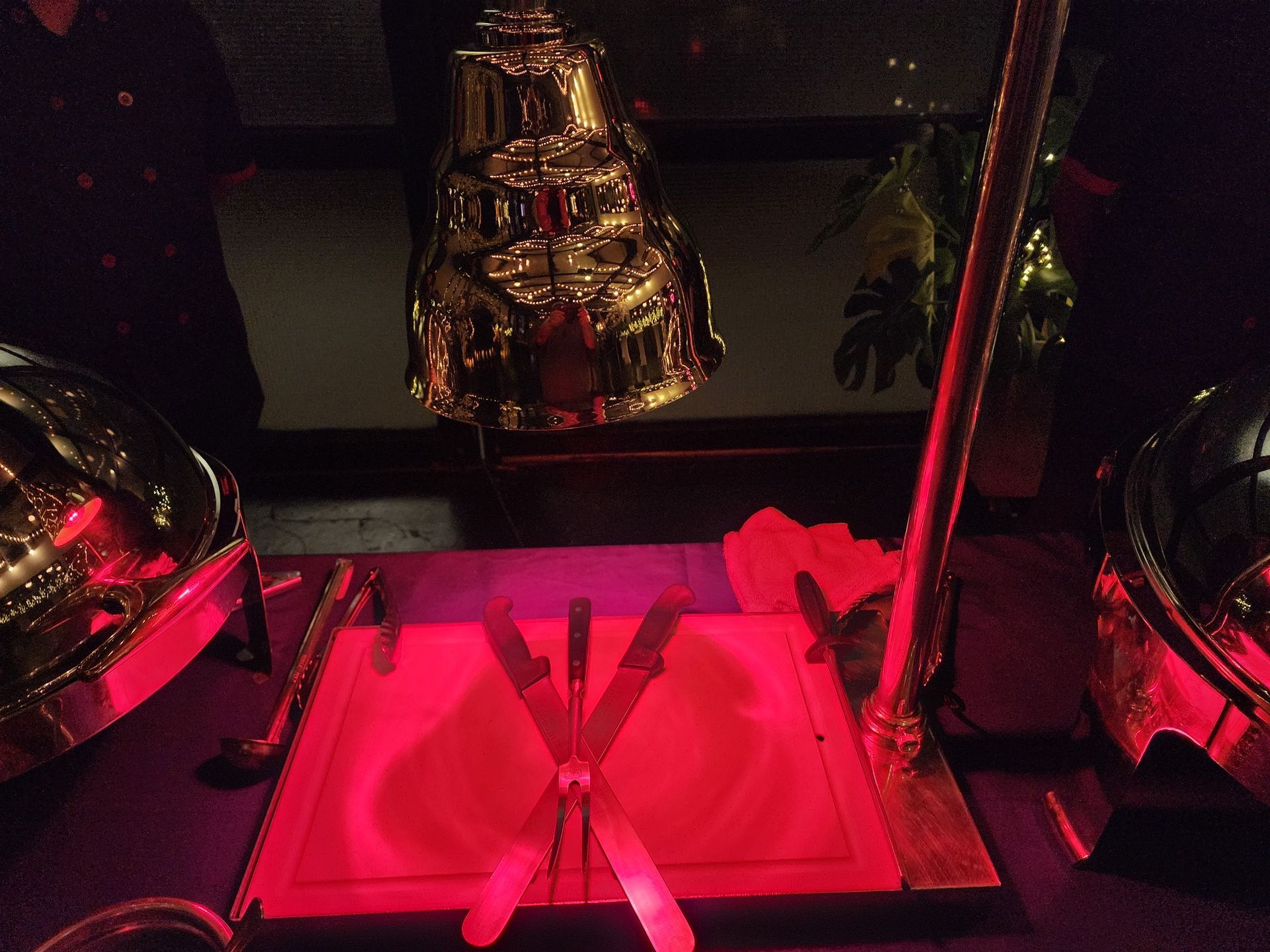 A food carving station with a heat lamp shining red light onto a cutting board with knives, flanked by two chafing dishes.
