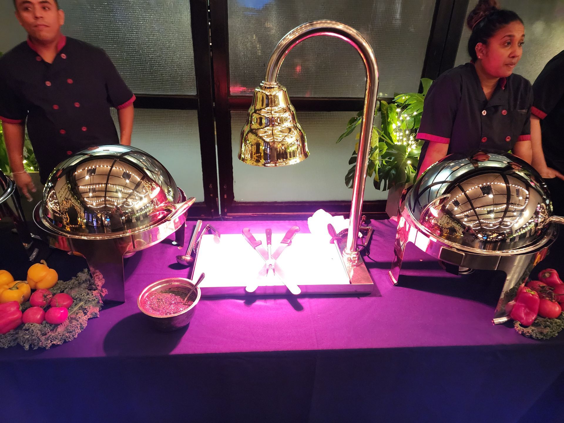 Two food servers stand behind a buffet table featuring two large silver chafing dishes and a gold-colored heat lamp.