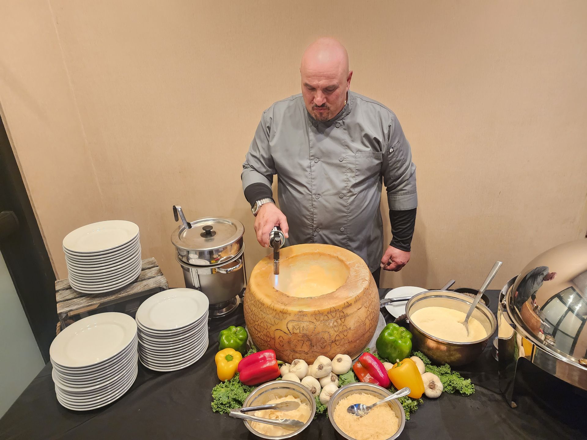 A chef prepares food in a large hollowed-out cheese wheel on a table with peppers, garlic, and serving bowls.