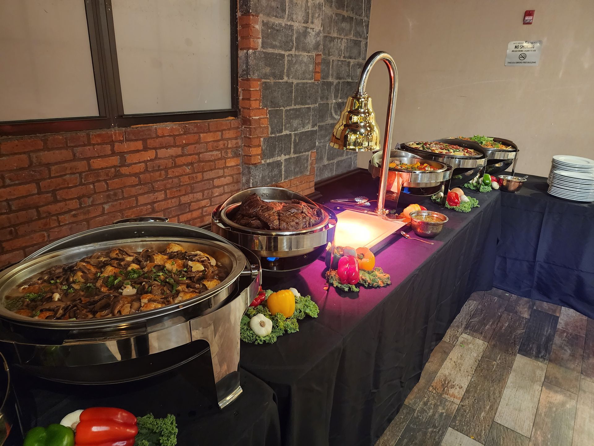 A buffet setup with silver chafing dishes filled with food on a black-clothed table against a brick wall.