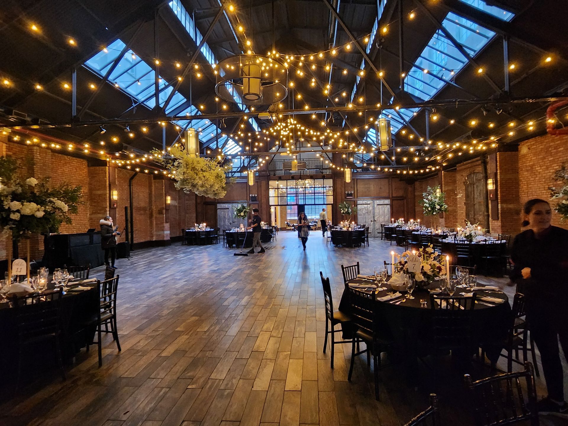 A rustic wedding venue with brick walls, wood floors, string lights, and round tables set for a reception.